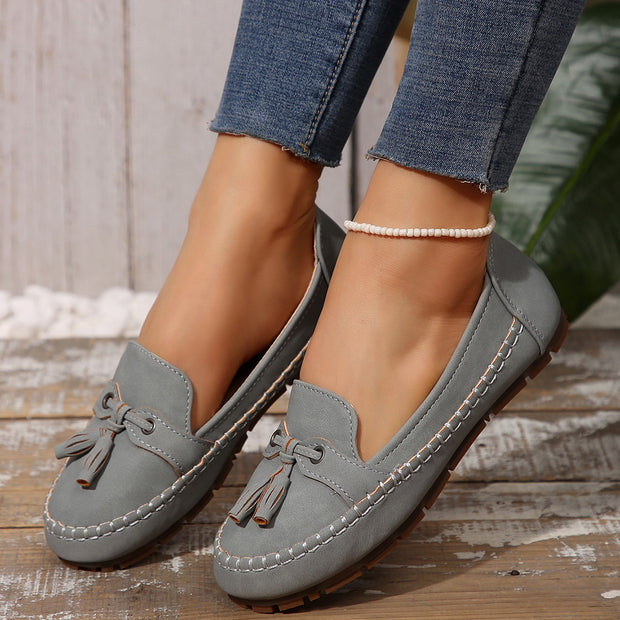 Gray loafers with tassels worn with blue jeans on a wooden floor.