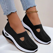 Black slip-on shoes with white soles worn with light blue jeans on a neutral background