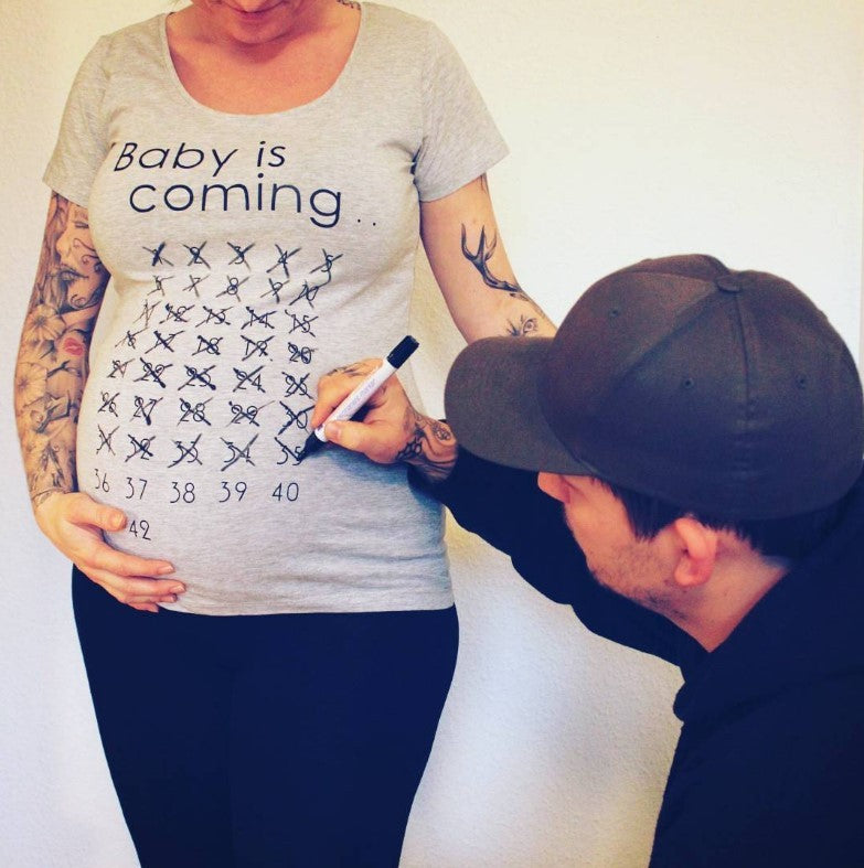 Pregnant person wearing a t-shirt with 'Baby is coming...' and a man writing on their belly.