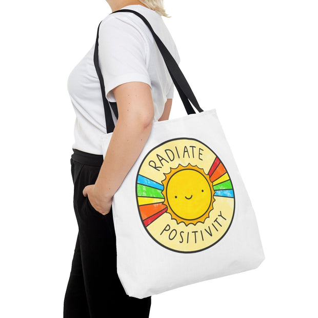Person holding a white tote bag with a sun design and 'Radiate Positivity' text.