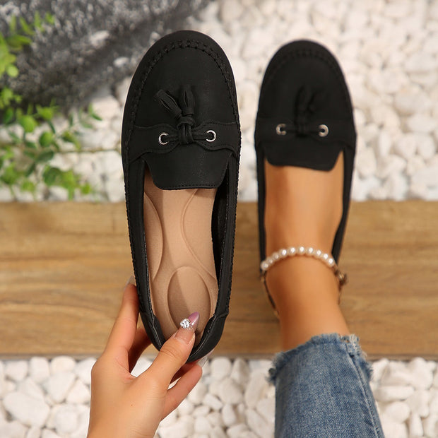 Black loafers with a bow held by a hand on a pebble surface.