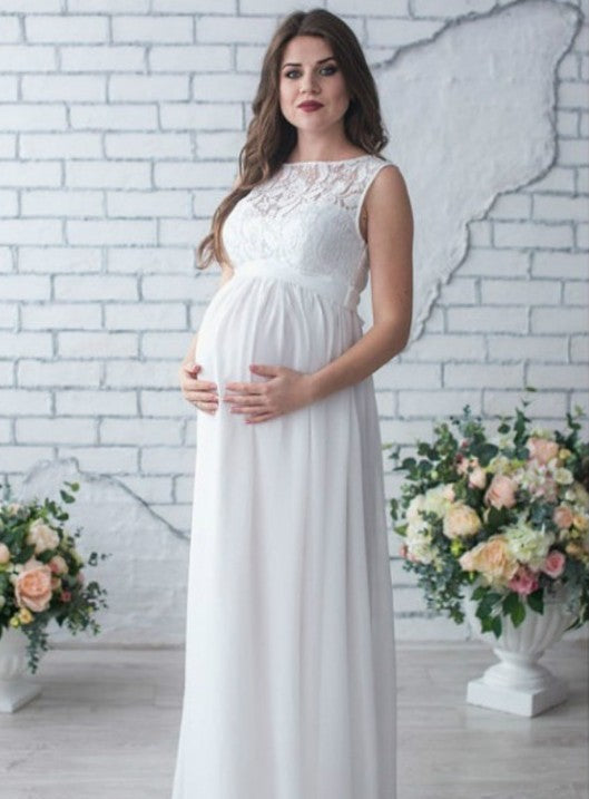 Woman in a white maternity dress standing in front of a white brick wall with floral decorations.