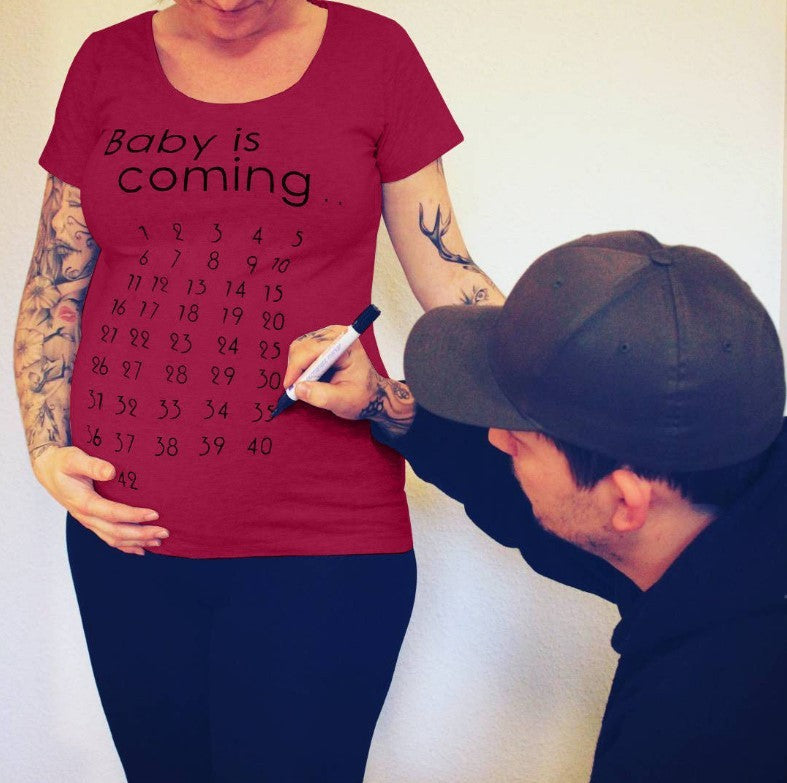 Pregnant person wearing a red t-shirt with 'Baby is coming...' and a countdown, being measured by a man.