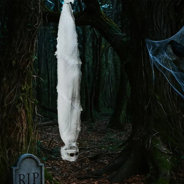Ghostly figure wrapped in white sheet hanging from a tree in a dark forest with tombstone.