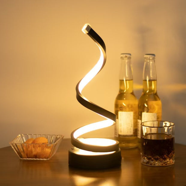 Spiral light fixture on a table with bottles and a glass of dark liquid.