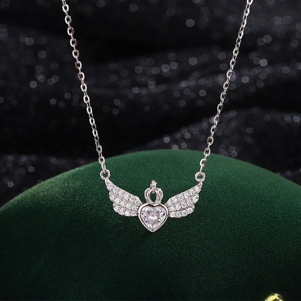 Silver necklace with angel wings and heart pendant on a green cushion against a dark background