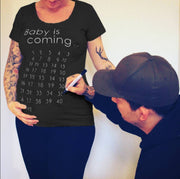 Person wearing a black t-shirt with 'Baby is coming' and pregnancy due date countdown, with another person marking the shirt.
