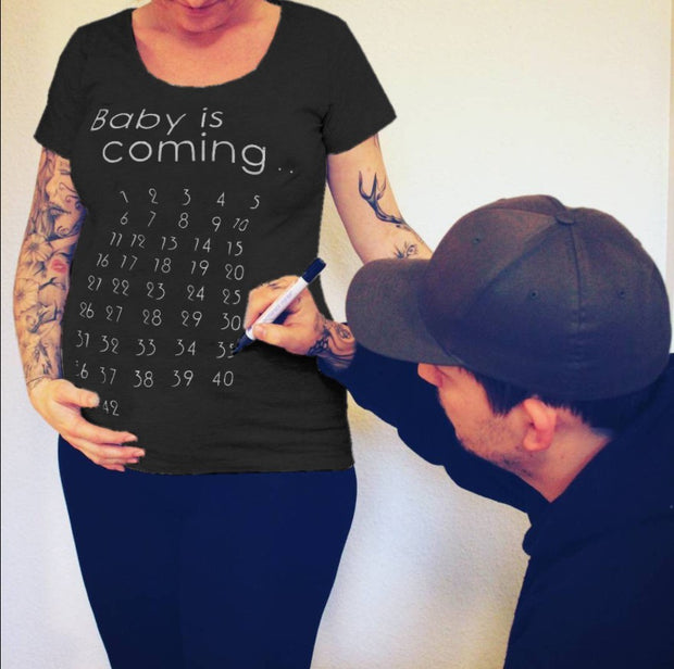 Person wearing a black t-shirt with 'Baby is coming' and pregnancy due date countdown, with another person marking the shirt.
