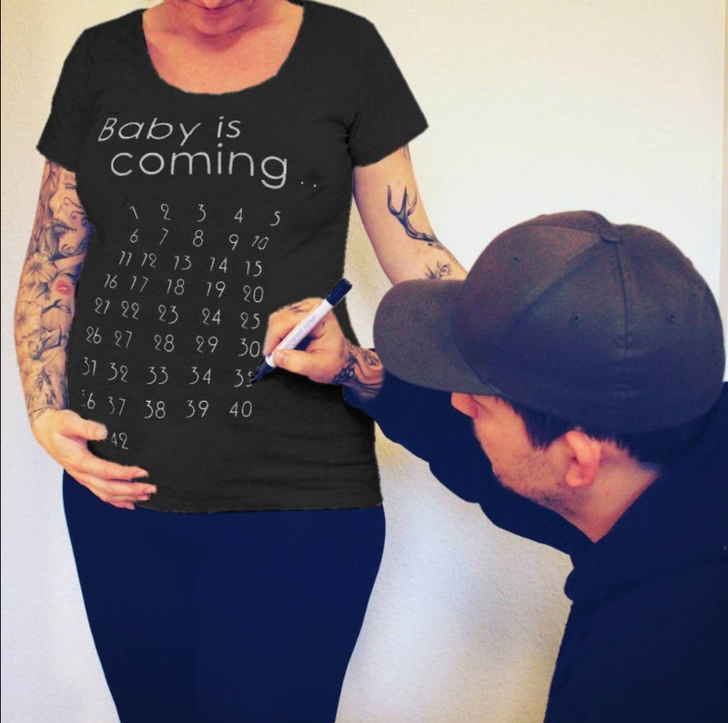 Person wearing a black t-shirt with 'Baby is coming' and pregnancy due date countdown, with another person marking the shirt.