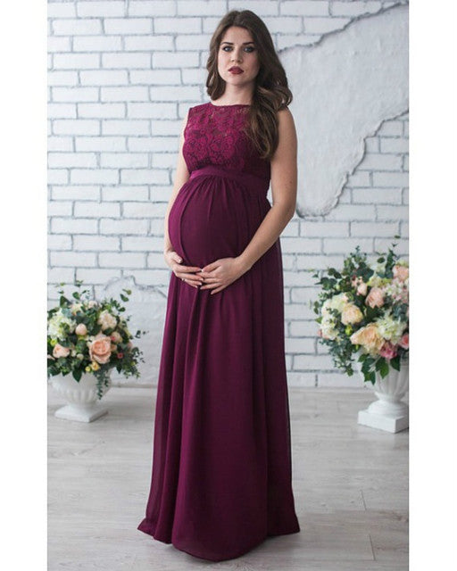 Woman wearing a burgundy maternity dress with floral lace top against a white brick wall.