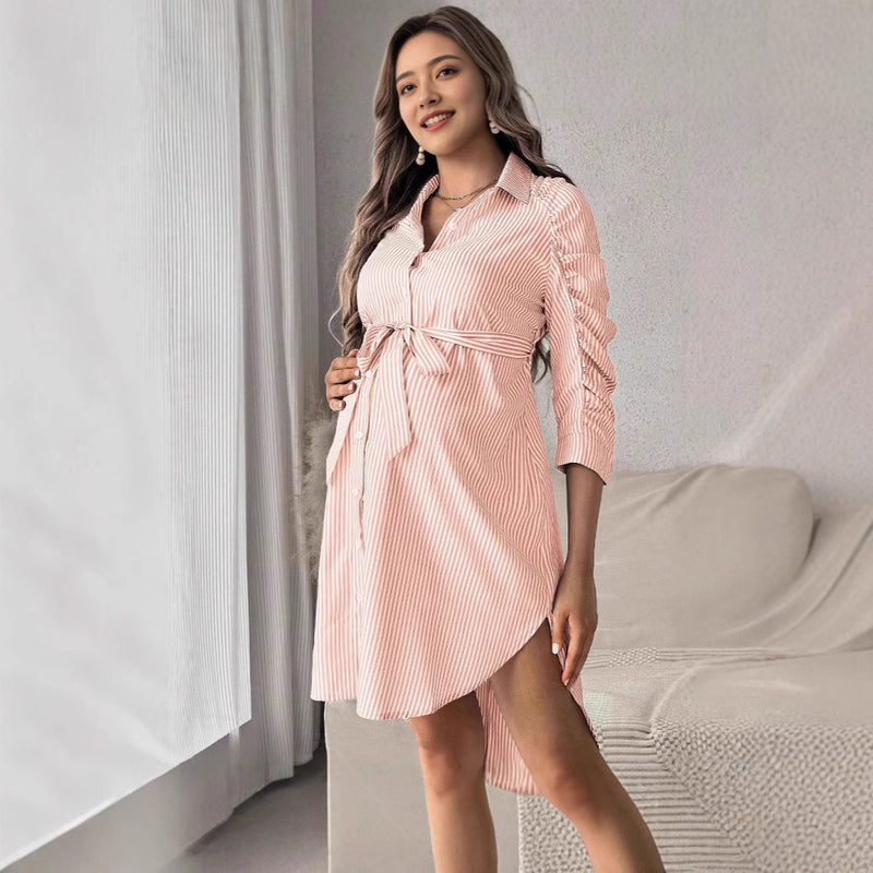 Woman wearing a pink striped dress standing in a room with a bed and curtains.