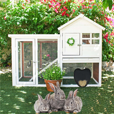 White rabbit hutch with a wreath and heart-shaped door sign, surrounded by greenery and three rabbits.
