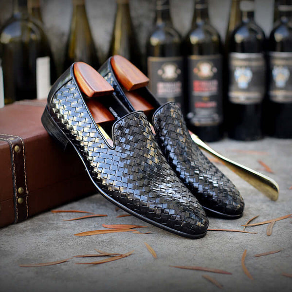 Black woven loafers with wooden soles on a textured surface with wine bottles in the background.