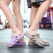 Two pairs of feet wearing purple and beige clogs on a blurred indoor background.