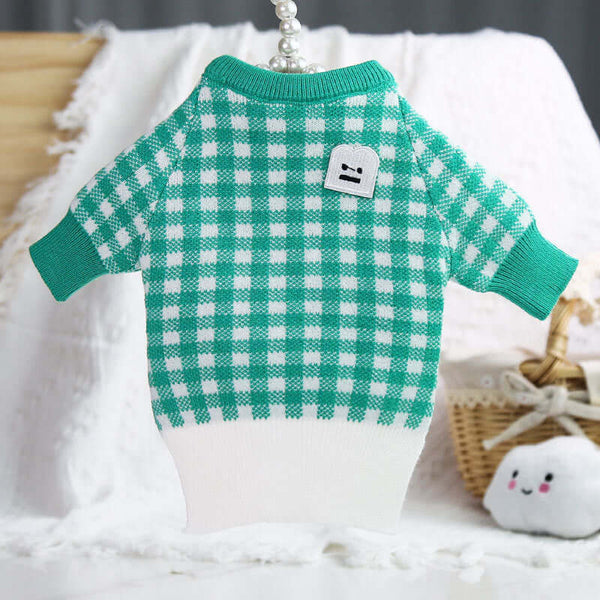 Green and white checkered dog sweater on a wooden surface with a soft white blanket and wicker basket in the background.
