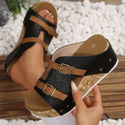 Black wedge sandals with brown straps and cork sole held by a hand on a pebbled surface.