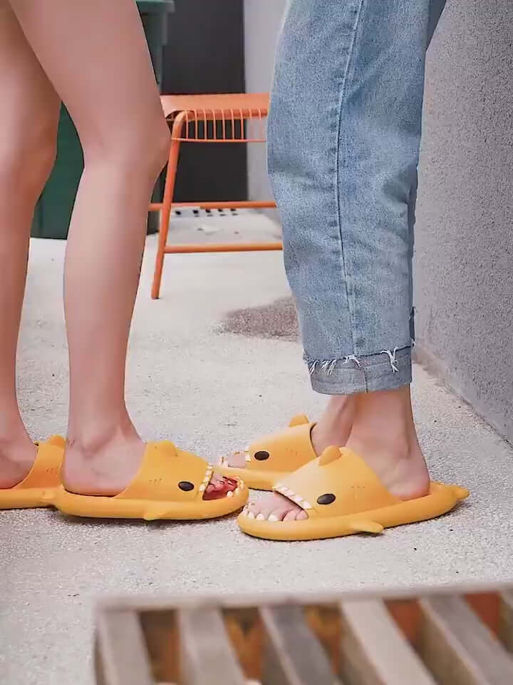 Pair of adults wearing cartoon shark slippers, showcasing bright yellow design and playful shark features.