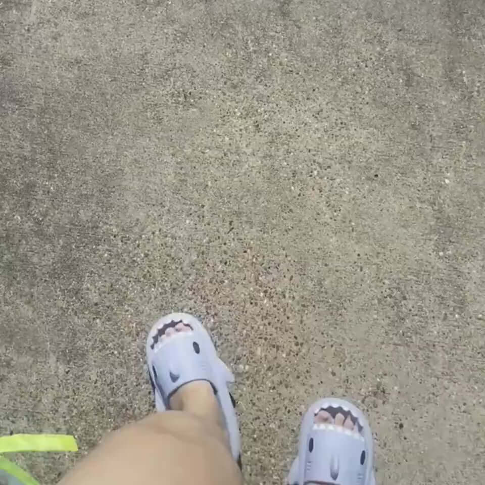 Adult wearing cartoon shark slippers walking on a pathway, showcasing fun indoor/outdoor design.